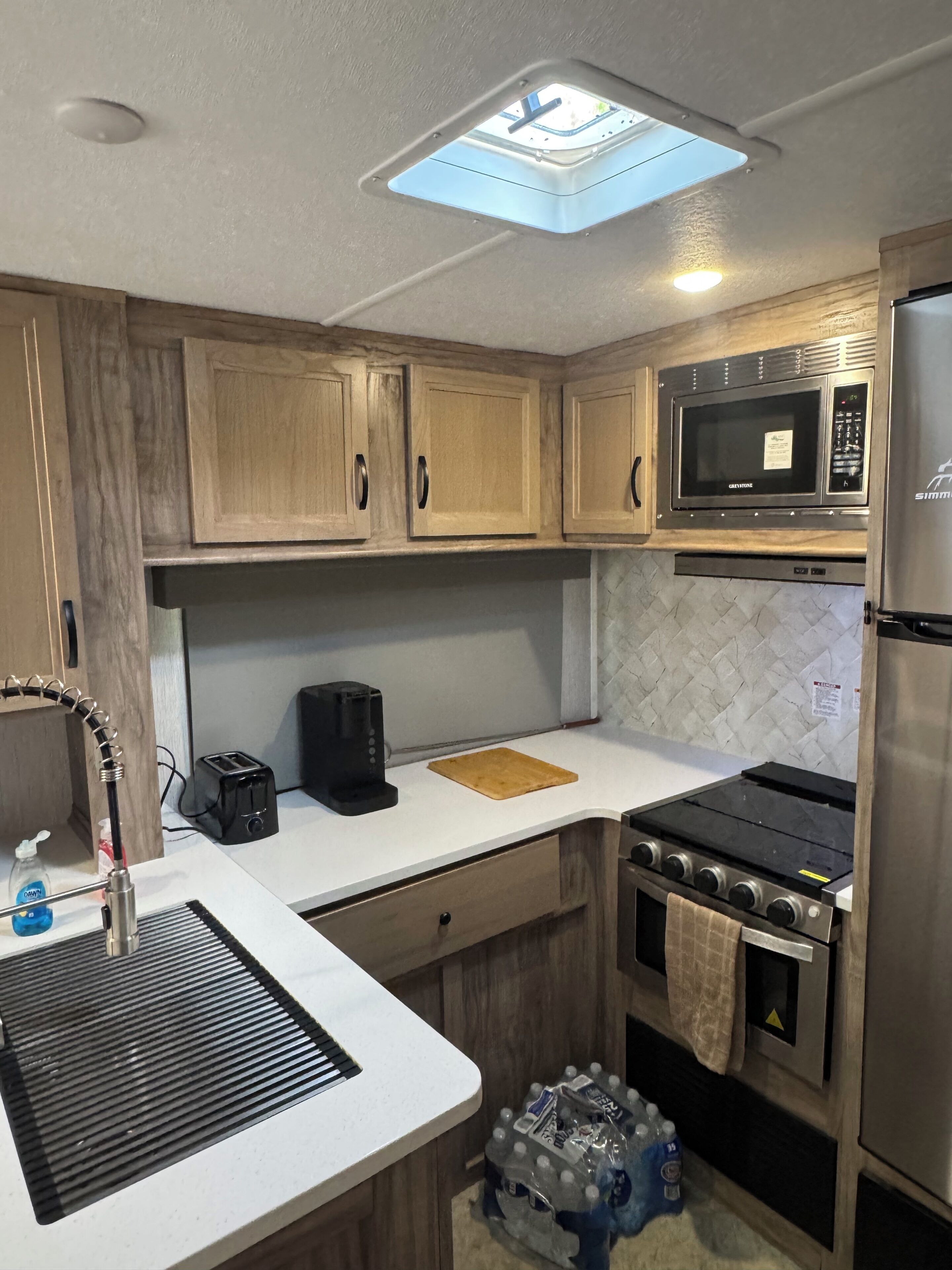 A compact kitchen with wooden cabinets, a microwave, a stove, a sink, and a skylight.