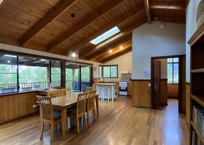 A spacious room with a wooden ceiling, a dining table with chairs, a kitchen area, and large windows offering views of trees.