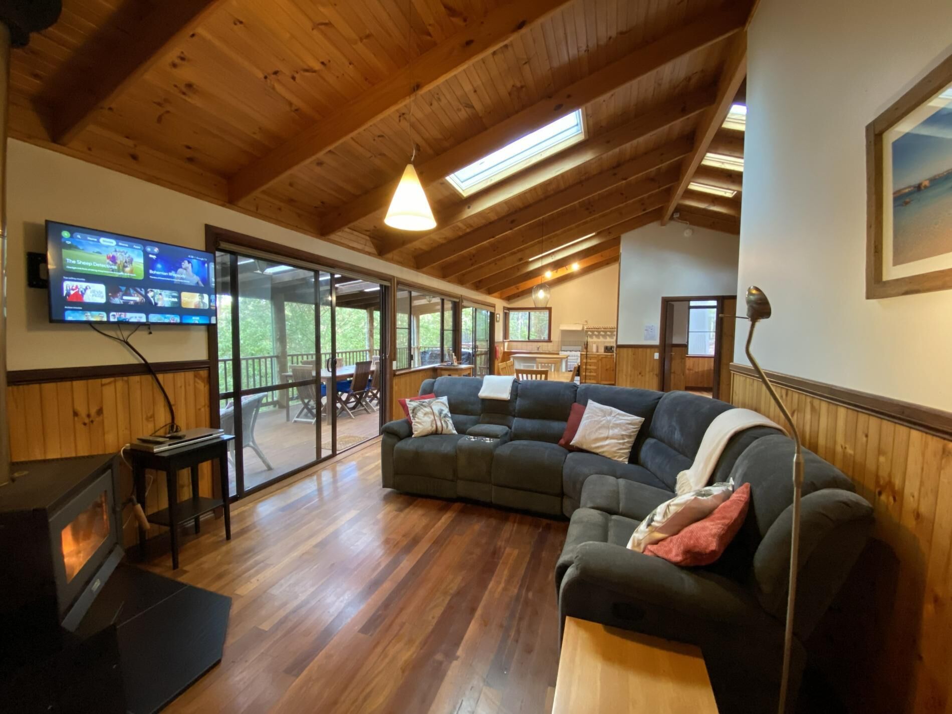 A cozy living room with a wooden ceiling, a flat-screen TV, a fireplace, and a large window overlooking a deck.