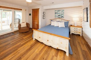 Ein Schlafzimmer mit einem hölzernen Bett, einem Sessel, einem Nachttisch und Blick ins Freie.