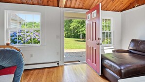 Ein Zimmer mit Holzboden, einer roten Tür, einem braunen Ledersofa und einem Fenster mit Gartenblick.
