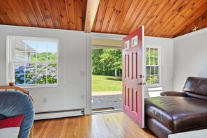 A room with a wooden ceiling, a red door, a brown leather sofa, and a window with a view of a garden. - Whispering Waters on Bearse Pond (Centerville)