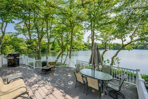 A deck with a table and chairs, a grill, and a view of a lake surrounded by trees. - Whispering Waters on Bearse Pond (Centerville)