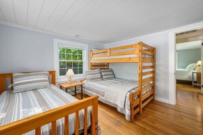 A room with two bunk beds, a small table with a lamp, and a window with a view of greenery. - Whispering Waters on Bearse Pond (Centerville)