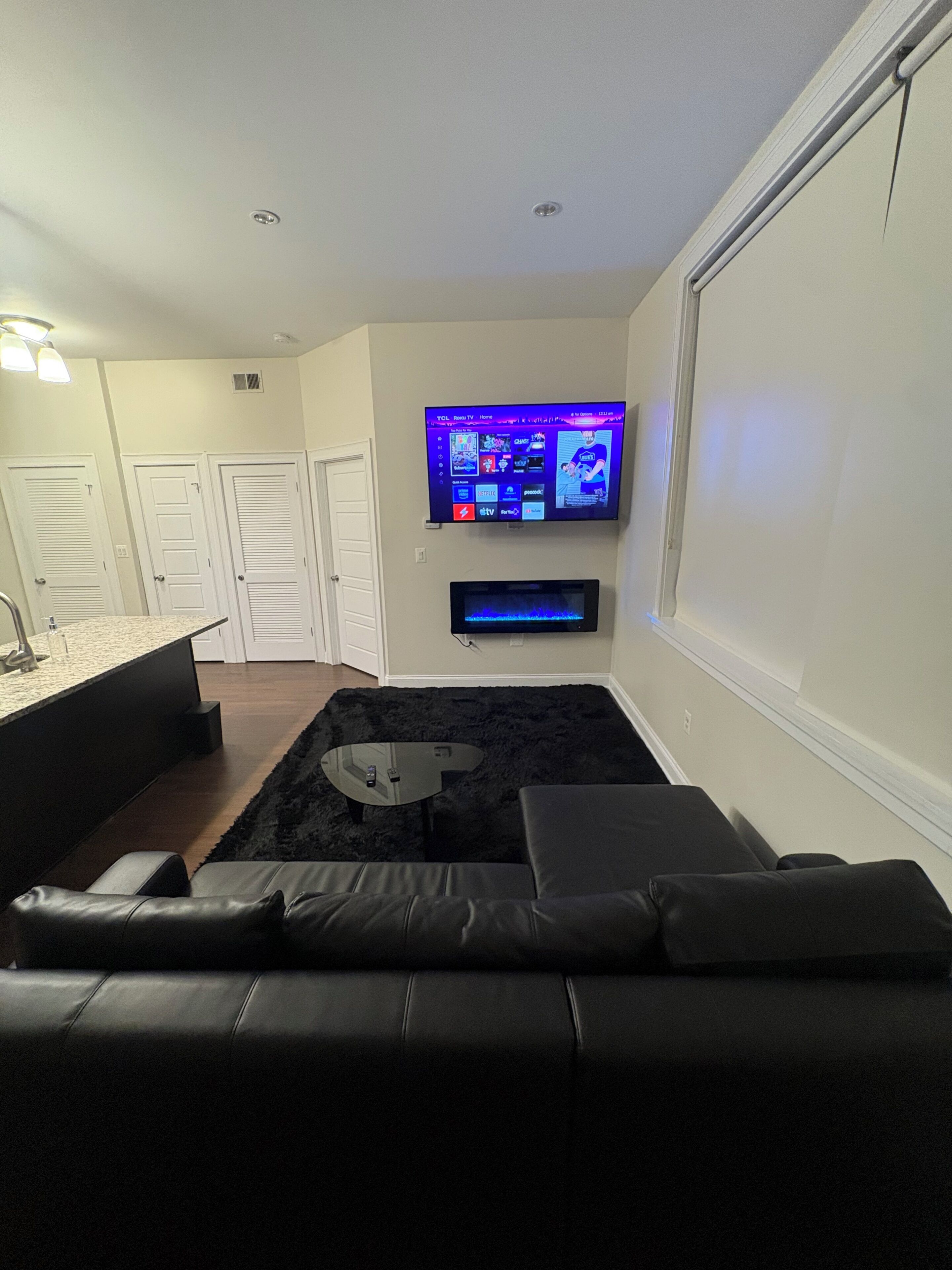 A modern living room with a flat-screen TV mounted on the wall, a black leather sofa, a glass coffee table, and a black rug.
