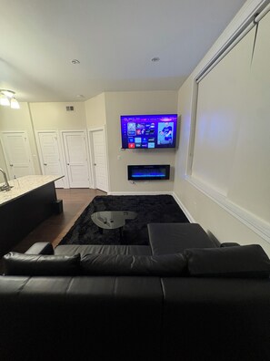 Ein modernes Wohnzimmer mit einem wandmontierten Flachbildfernseher, einem schwarzen Ledersofa, einem Glastisch und einem schwarzen Teppich.