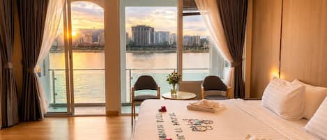 Habitación de hotel con una cama grande, vistas de la ciudad a través del ventanal y un balcón con mesa y sillas.