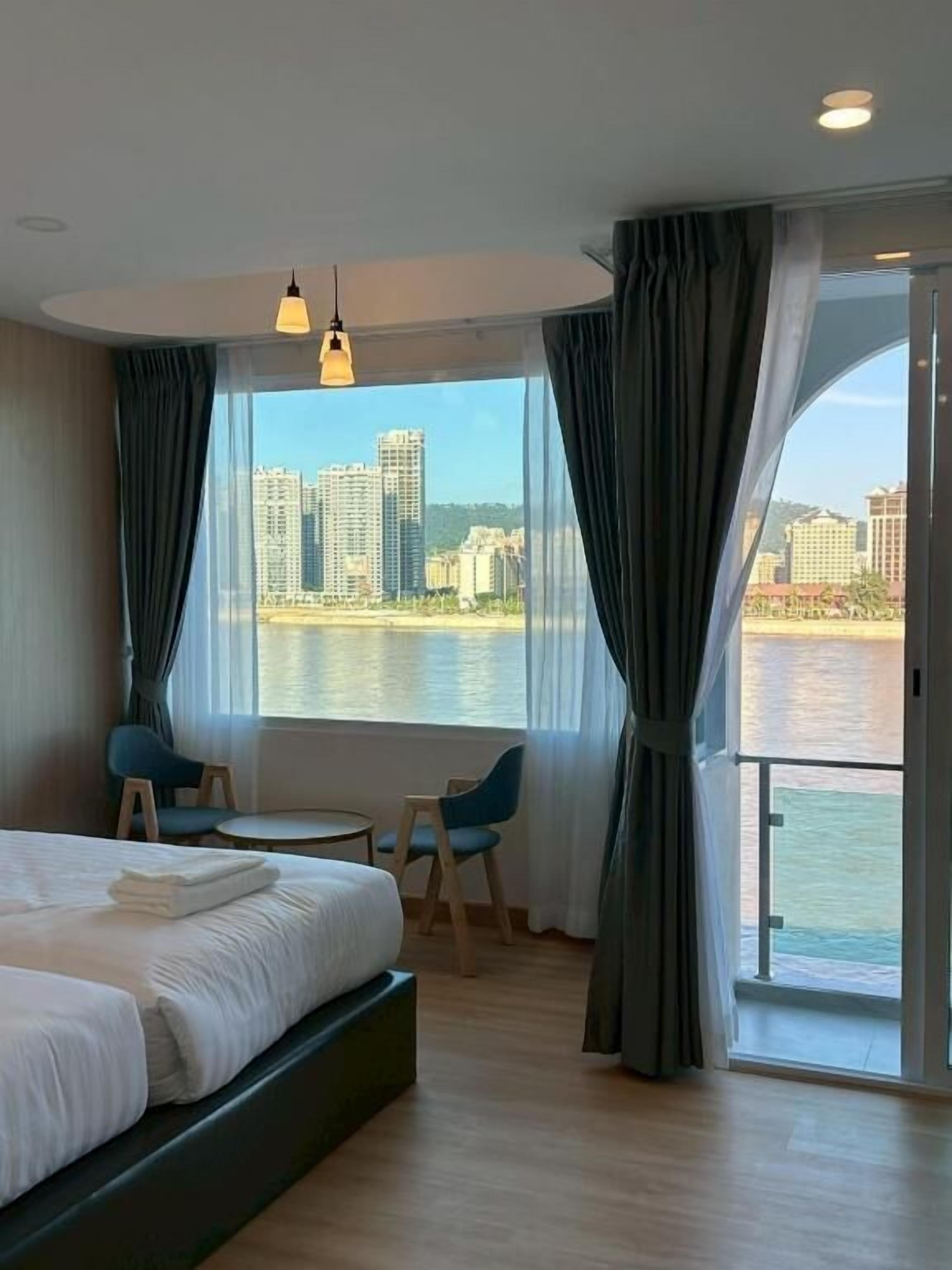 Habitación de hotel con cama, una mesita, dos sillas y un ventanal que ofrece vistas a un cuerpo de agua y edificios.