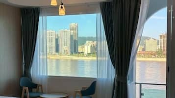 Habitación de hotel con cama, una mesita, dos sillas y un ventanal que ofrece vistas a un cuerpo de agua y edificios.