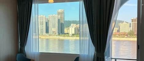 A hotel room with a bed, a small table, two chairs, and a large window offering a view of a body of water and buildings.