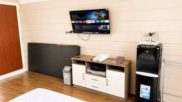 A hotel room with a bed, a TV mounted on the wall, a small wooden desk, a water dispenser, and a trash bin.