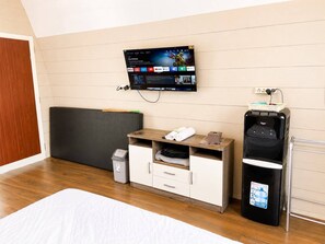 A hotel room with a bed, a TV mounted on the wall, a small wooden desk, a water dispenser, and a trash bin.
