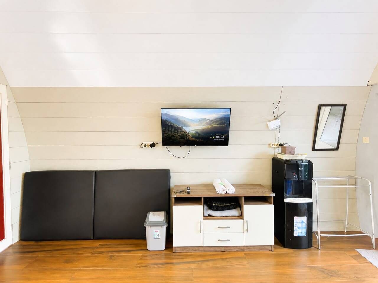 Una habitación con piso de madera, un sofá negro, un televisor montado en la pared, un dispensador de agua y un pequeño cubo de basura.