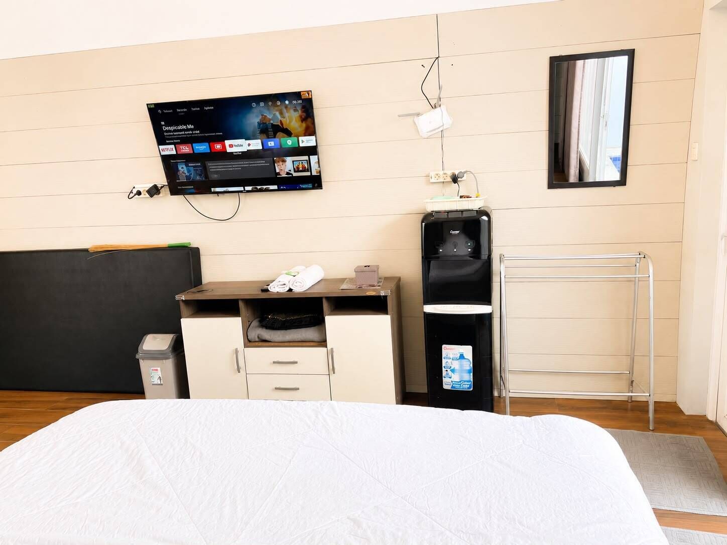 A hotel room with a bed, a desk with a computer, a TV mounted on the wall, a water dispenser, and a towel rack.