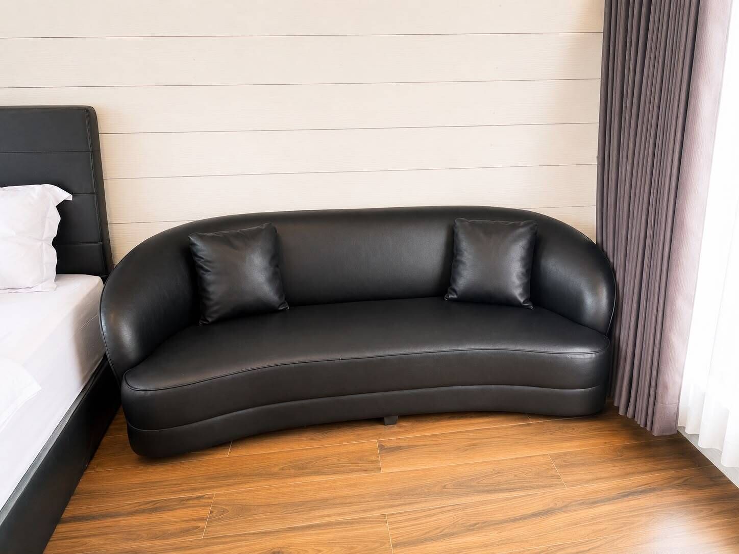 A black leather sofa against a wooden floor, a white bed with a white pillow, and a curtain.