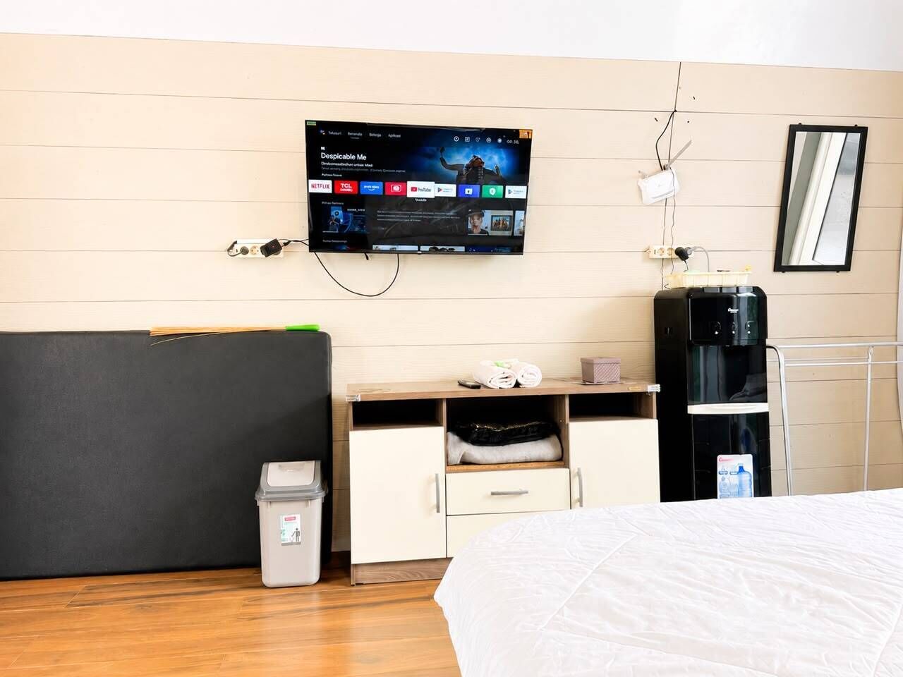 Habitación de hotel con cama, escritorio con monitor de computadora, dispensador de agua y basurero.