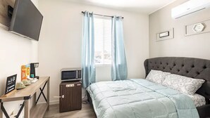 A bedroom with a bed, a TV mounted on the wall, a desk with a microwave, and a window with curtains.