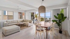 A spacious living room with a large sofa, a dining table with chairs, and a large plant. - 3/39 Albatross Avenue (Mermaid Beach)