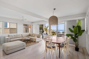 A spacious living room with a large sofa, a dining table with chairs, and a large plant. - 3/39 Albatross Avenue (Mermaid Beach)