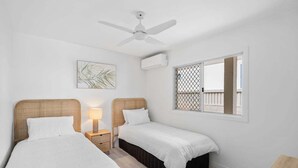 A bedroom with two single beds, a ceiling fan, an air conditioning unit, a window with a view, and a small bedside table with a lamp. - 3/39 Albatross Avenue (Mermaid Beach)