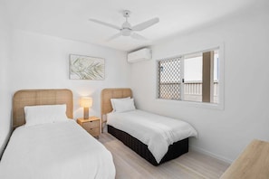 A bedroom with two single beds, a ceiling fan, an air conditioning unit, a window with a view, and a small bedside table with a lamp. - 3/39 Albatross Avenue (Mermaid Beach)