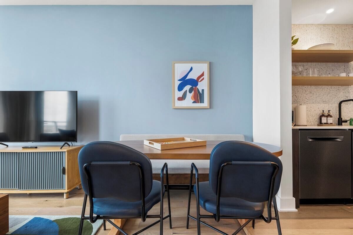Una sala de estar moderna con una televisión de pantalla plana, dos sillas azules, una mesa de comedor de madera y una cocina con estantes y lavavajillas.