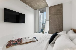 A modern hotel room with a large bed, a flat-screen TV mounted on the wall, and a view of the city through a large window. - High Rise Suite with a View | Rainey Street Fun & Entertainment! (Austin)