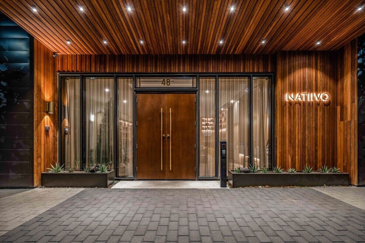 A modern building entrance with a wooden facade, a large glass door, and the name 'NATIVO' illuminated on the right.