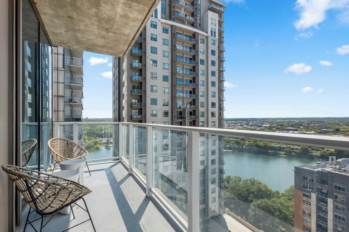 A balcony with wicker chairs, a glass railing, and a view of a river and buildings.
