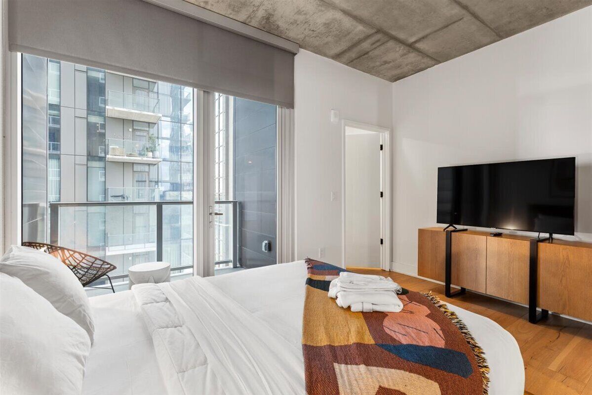Habitación de hotel con cama, televisor, una mesita y vistas a un edificio a través de amplias ventanas.