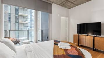 Habitación de hotel con cama, televisor, una mesita y vistas a un edificio a través de amplias ventanas.