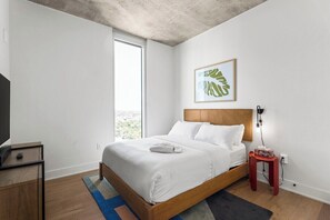 A bedroom with a bed, a television, a nightstand, a red side table, and a window with a view. - 2601 ATX Skyline Corner Retreat | Rooftop Pool, Skyline Views, Rainey St (Austin)