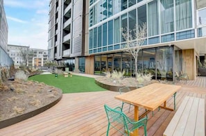 A modern outdoor patio with a wooden table and chairs, surrounded by a landscaped garden and a building with large windows. - Luxurious Executive Condo | Downtown High-Rise + *Rooftop Pool* (Austin)