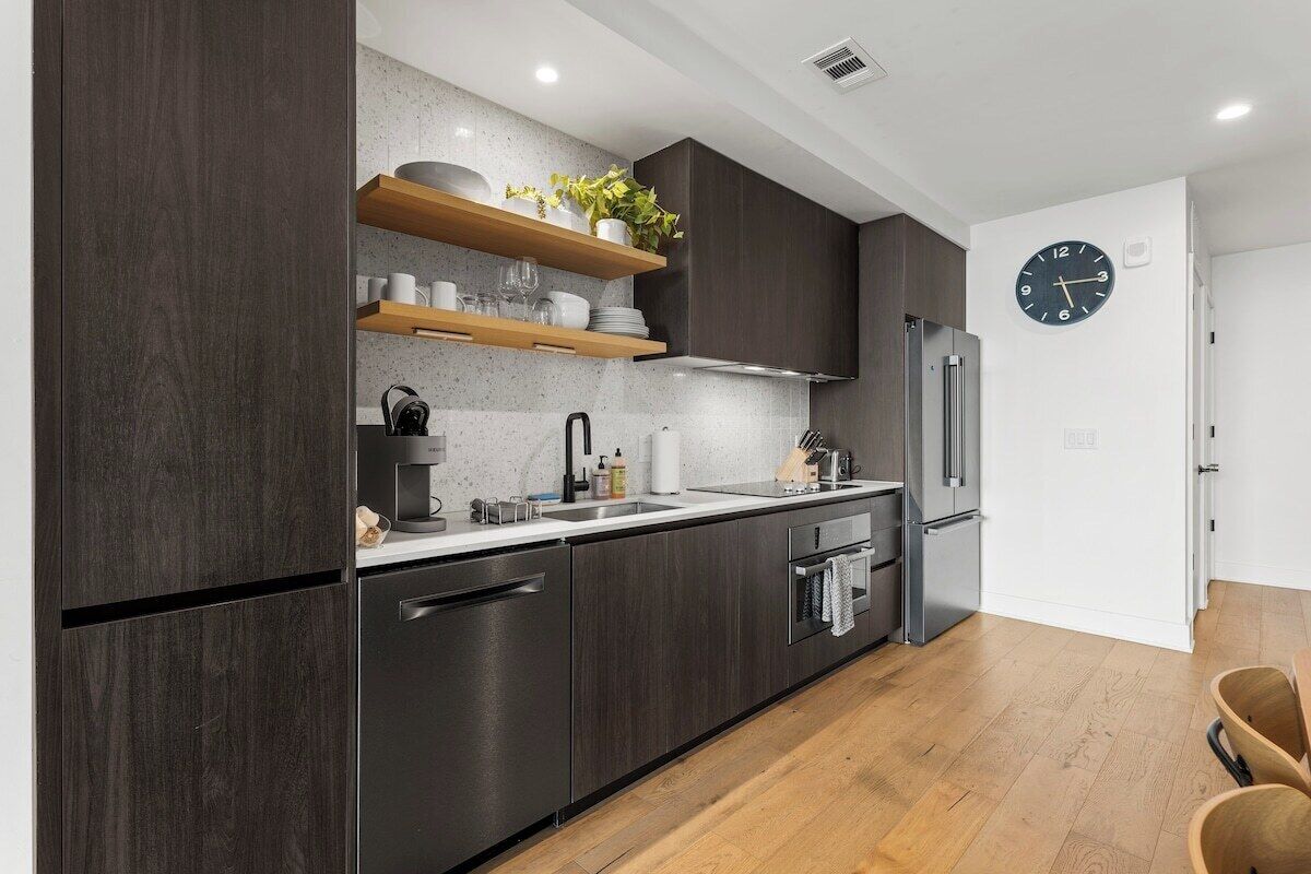 A modern kitchen with dark cabinetry, stainless steel appliances, wooden shelves, and a wall clock.
