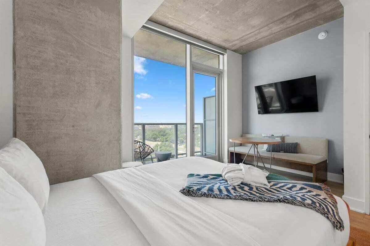 Una habitación de hotel moderna con una cama grande, un televisor montado en la pared, un balcón con vistas y una pequeña mesa con una silla.