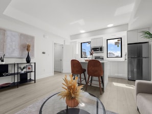 A modern living room with a glass coffee table, a sofa, and a kitchen with white cabinets and a stainless steel refrigerator. - Cozy 2BR Penthouse Apt with Rooftop and Parking, Near to Falls (Niagara Falls)