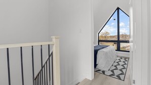 Uma escada estreita leva a um quarto com uma cama, uma janela e um tapete com padrão geométrico.