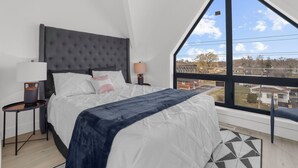 Quarto com uma cama grande, uma mesa de cabeceira, uma luminária e vista para uma área residencial pela janela.