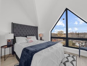 Ein Schlafzimmer mit einem großen Bett, einem Nachttisch, einer Lampe und Blick auf eine Wohngegend durch ein großes Fenster.