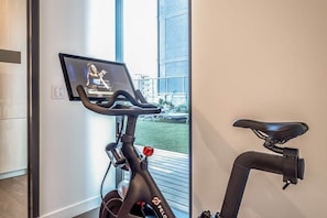 An indoor exercise bike with a screen displaying a person exercising, a view of a building outside, and a door to the left. - Deluxe Condo | Cool Vibes & Rooftop Pool! (Austin)