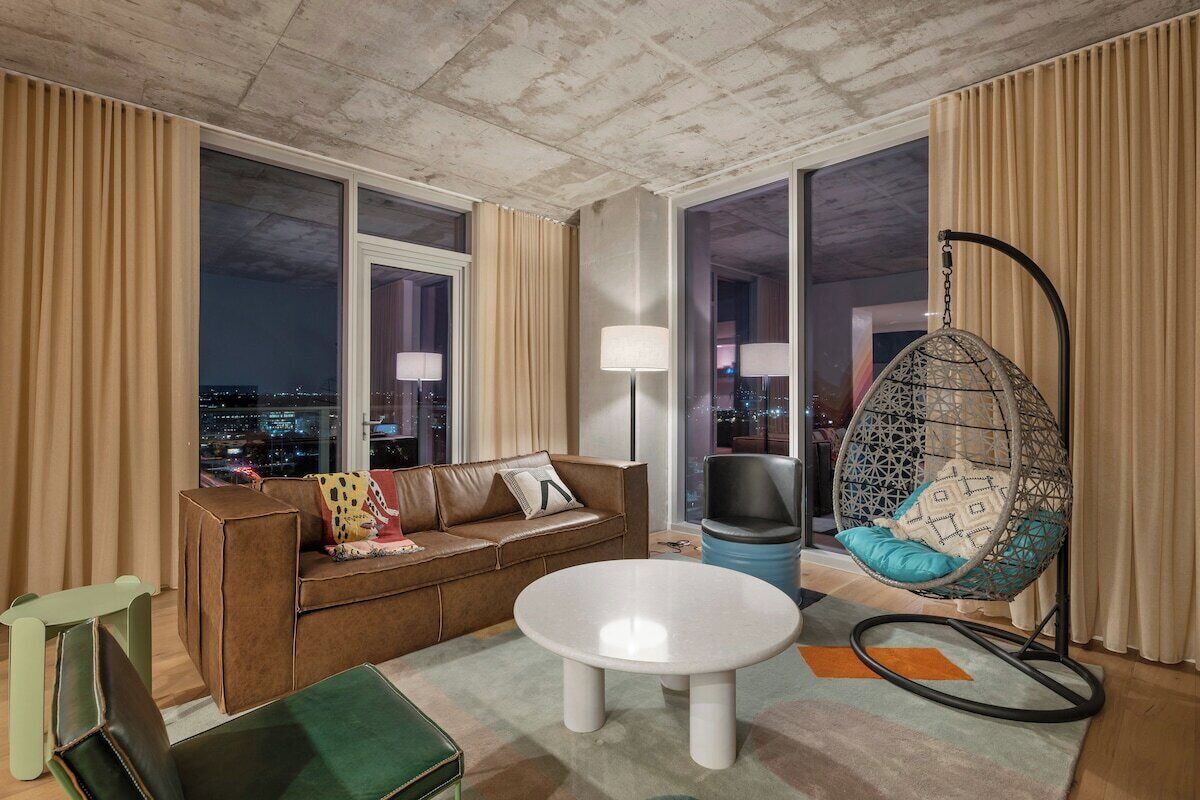 A modern living room with a sofa, a round coffee table, a hanging chair, and large windows offering a city view.