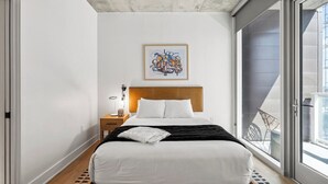 A modern bedroom with a wooden bed, white bedding, a framed abstract painting, a nightstand with a lamp, and a balcony view. - Riverside Penthouse in heart of Rainey - Sleeps 6 (Austin)