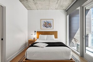 Ein modernes Schlafzimmer mit einem hölzernen Bett, weißer Bettwäsche, einem gerahmten abstrakten Gemälde, einem Nachttisch mit Lampe und Blick auf einen Balkon.