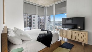 A modern bedroom with a large bed, a television on a wooden console, and a city view through large windows. - Riverside Penthouse in heart of Rainey - Sleeps 6 (Austin)