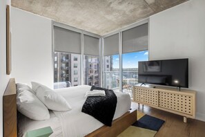 Ein modernes Schlafzimmer mit einem großen Bett, einem Fernseher auf einer Holzkonsole und Blick auf die Stadt durch große Fenster.