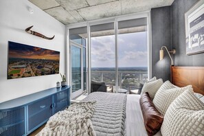 Ein Schlafzimmer mit einem großen Bett, einem Stadtbild, einer blauen Konsole und Blick auf die Stadt durch große Fenster.