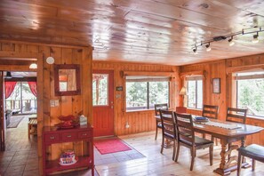 A wooden cabin interior with a dining table, chairs, a red door, and large windows.