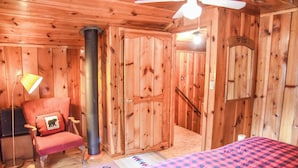 Interior de uma cabana de madeira com uma cama, uma cadeira, um abajur e um ventilador de teto.