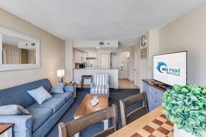 A furnished living room with a sofa, coffee table, and chairs, a kitchen area with cabinets and appliances, and a television displaying 'BIG FISH RENTALS'. - Amazing Oceanfront Views at Carolinian with Pool (Myrtle Beach)