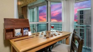 A wooden table with chess pieces and a glass of red wine, set against a window with a view of a sunset. - Amazing Oceanfront Views at Carolinian with Pool (Myrtle Beach)